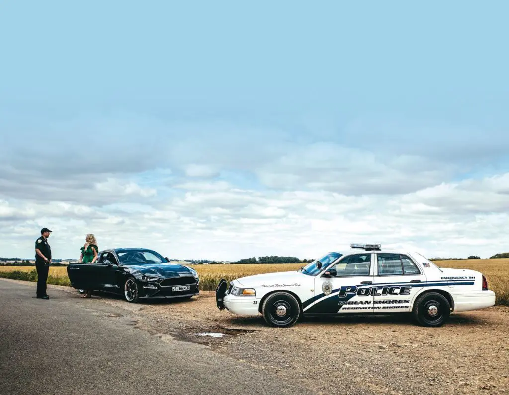 Ewan's Crown Victoria police car parked on the side of the road as Ewan in an American police outfit has a woman step out of her car.