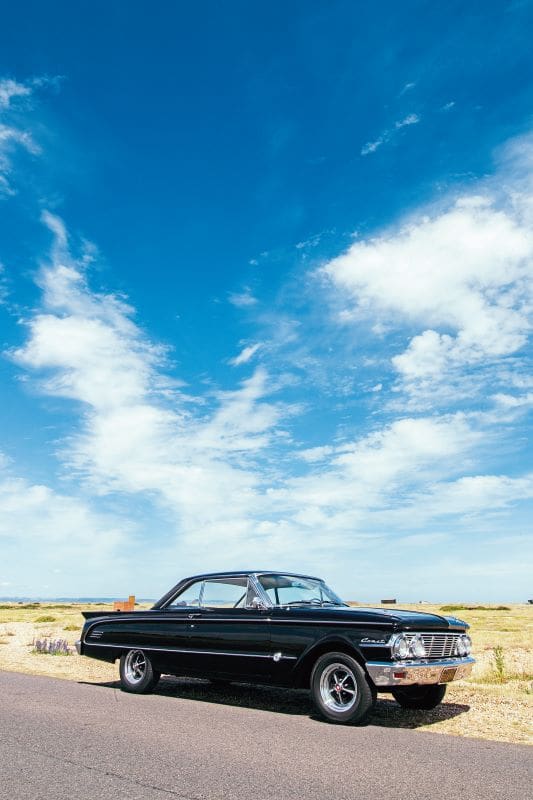 Full shot of the black Mercury Comet parked on the side of the road against a blue sky