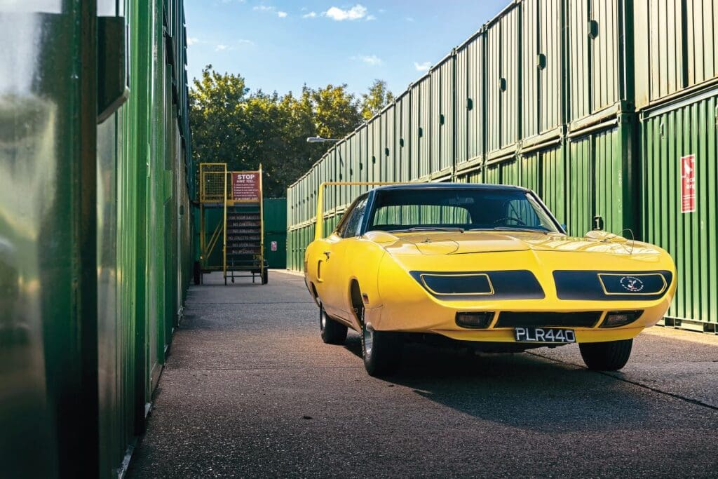 Bright yellow 1970 Plymouth Superbird parked among green shipping containers
